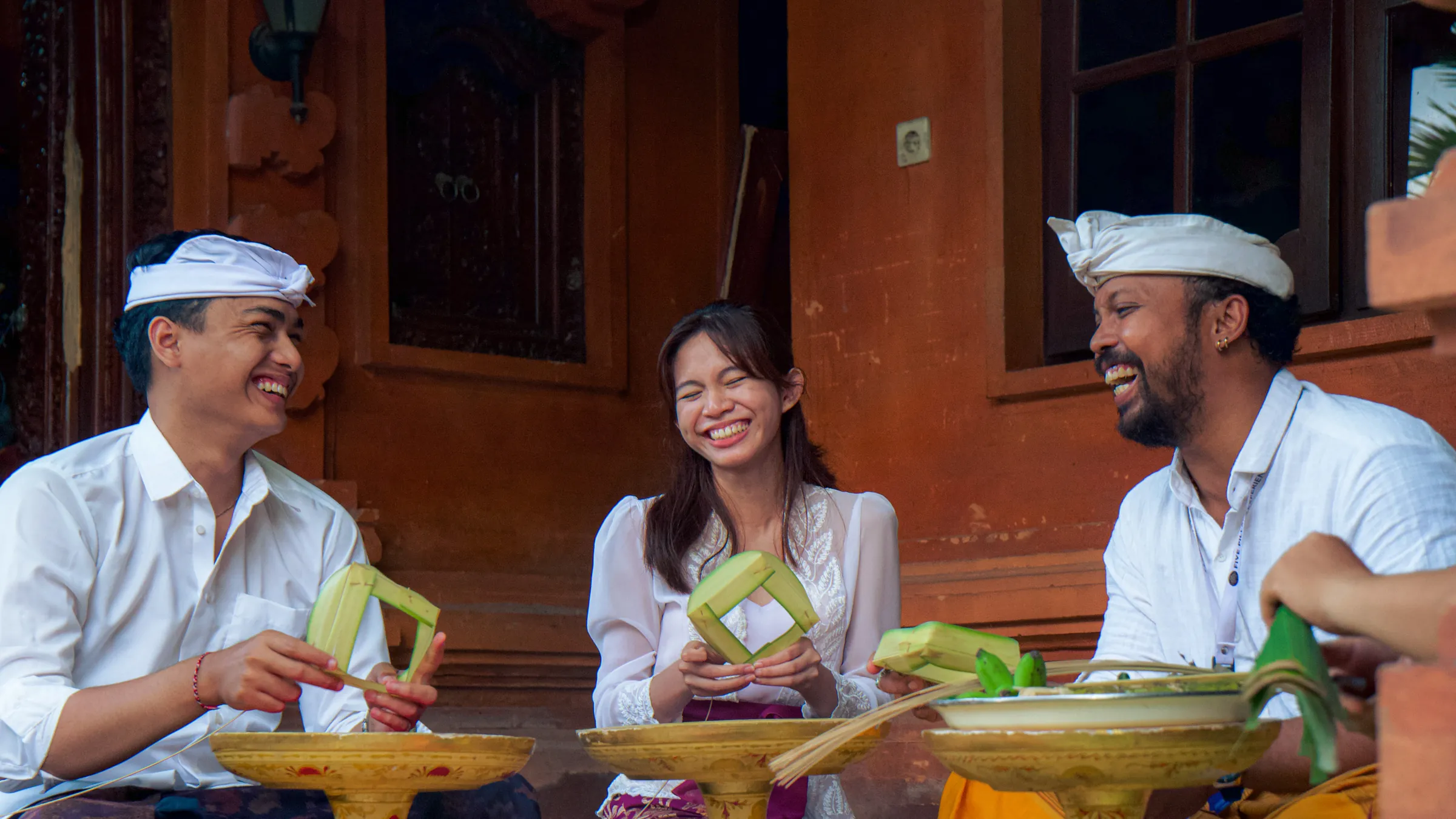 Two travelers laughing with a local host while folding palm-leaf offerings in a Balinese family courtyard