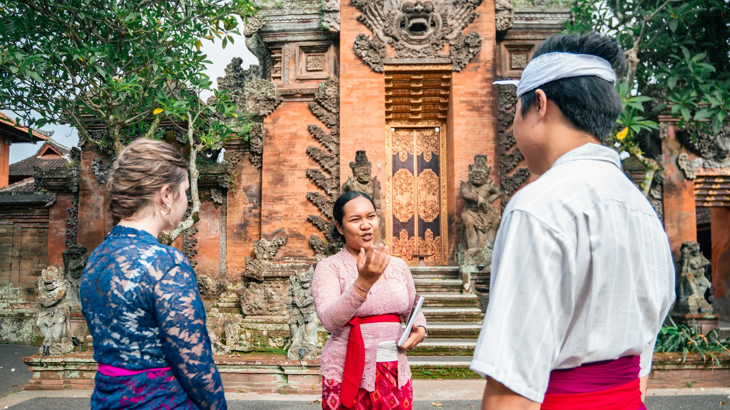 A local guide telling the story of a Balinese palace gate to two travelers in traditional dress